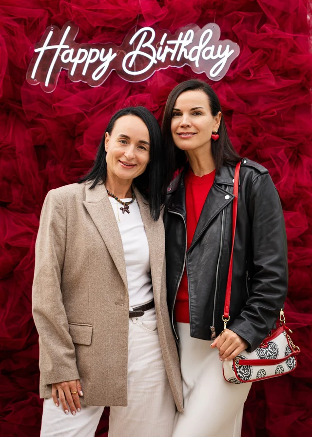 Two women in front of a red floral backdrop with an illuminated Happy Birthday sign