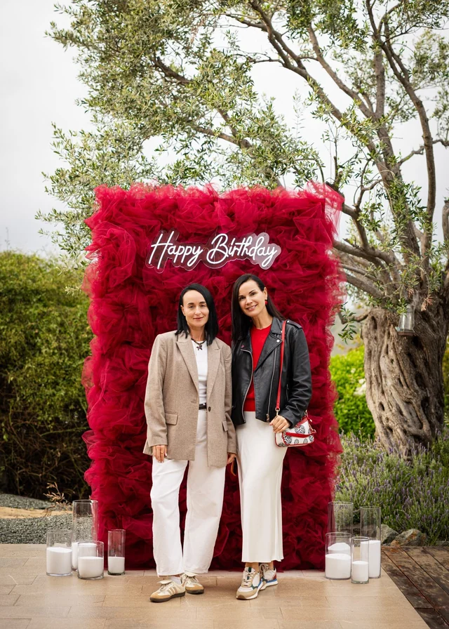 Two women standing beside candles in front of a red birthday backdrop beneath trees