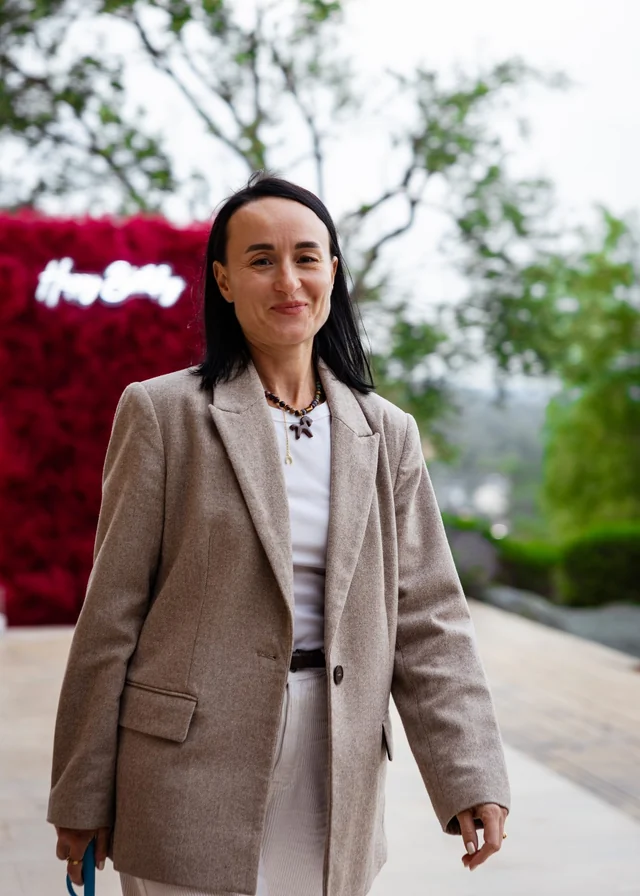 A woman in a beige jacket posing outdoors among trees