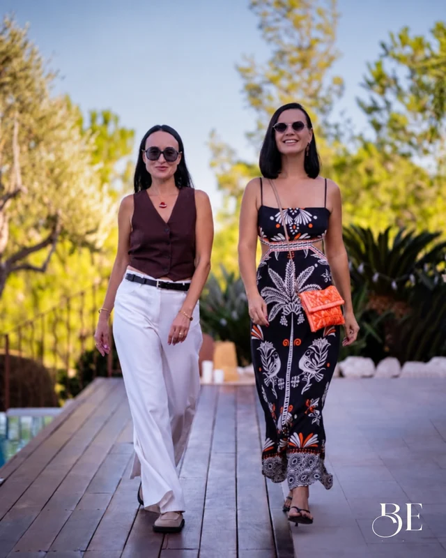 Two women wearing sunglasses walking along a wooden terrace, with the BE logo in the corner