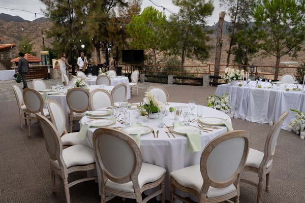 A round wedding table with white flowers and upholstered chairs beneath the trees