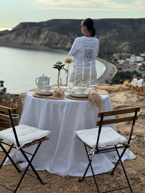 A woman standing beside the picnic table with the coast behind her