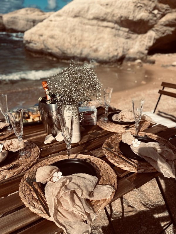 A picnic table with woven placemats and glassware beside the sea