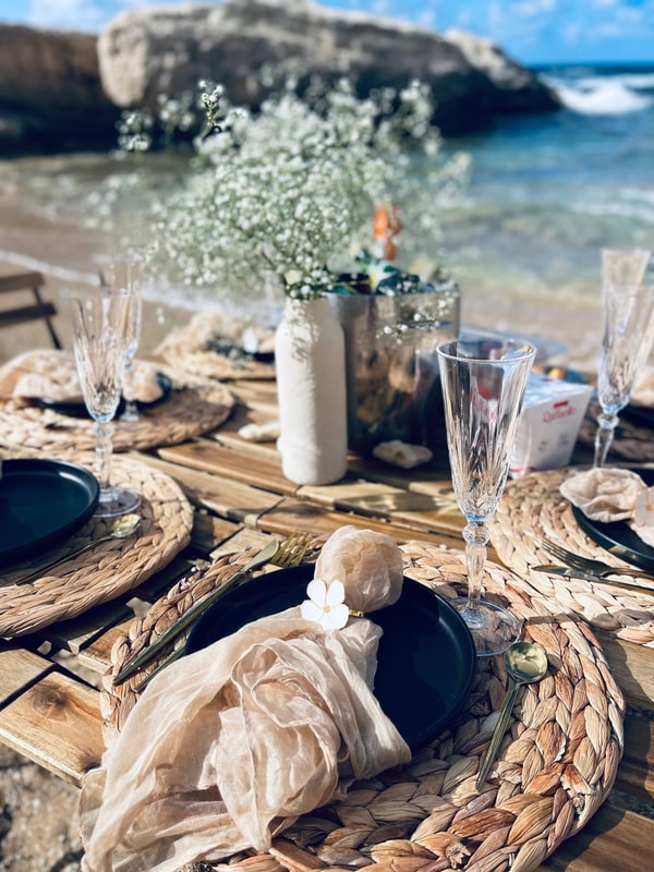 A picnic table with woven placemats and glassware beside the sea