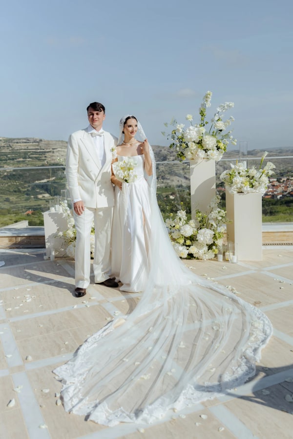 Lesya and Nikita in white beside the wedding flowers, with hills behind them