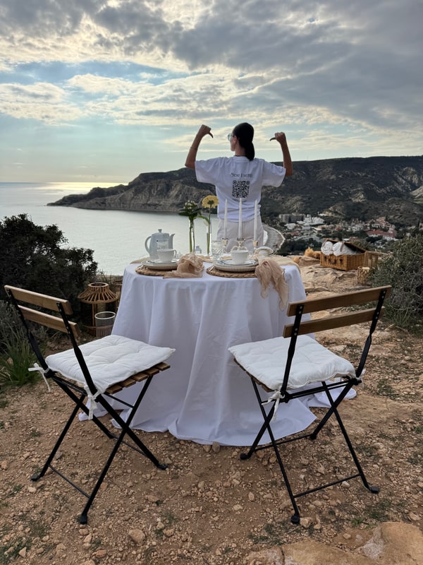 A woman standing beside the picnic table with the coast behind her