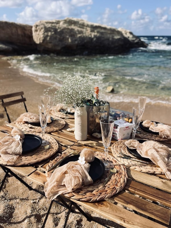 A picnic table with woven placemats and glassware beside the sea