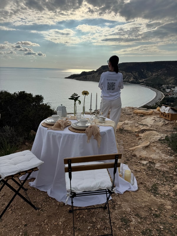 A woman standing beside the picnic table with the coast behind her