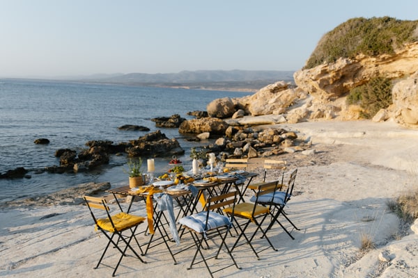 A long picnic table with yellow chairs on the rocky shore