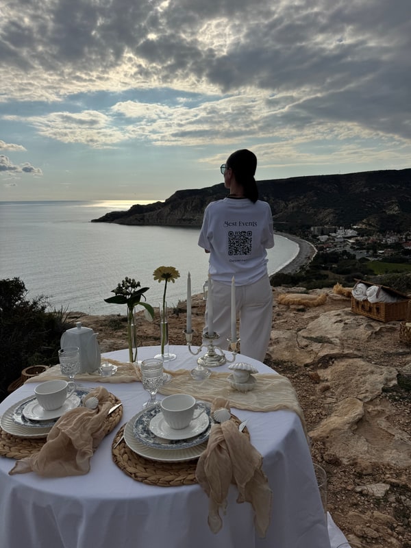 A woman standing beside the picnic table with the coast behind her