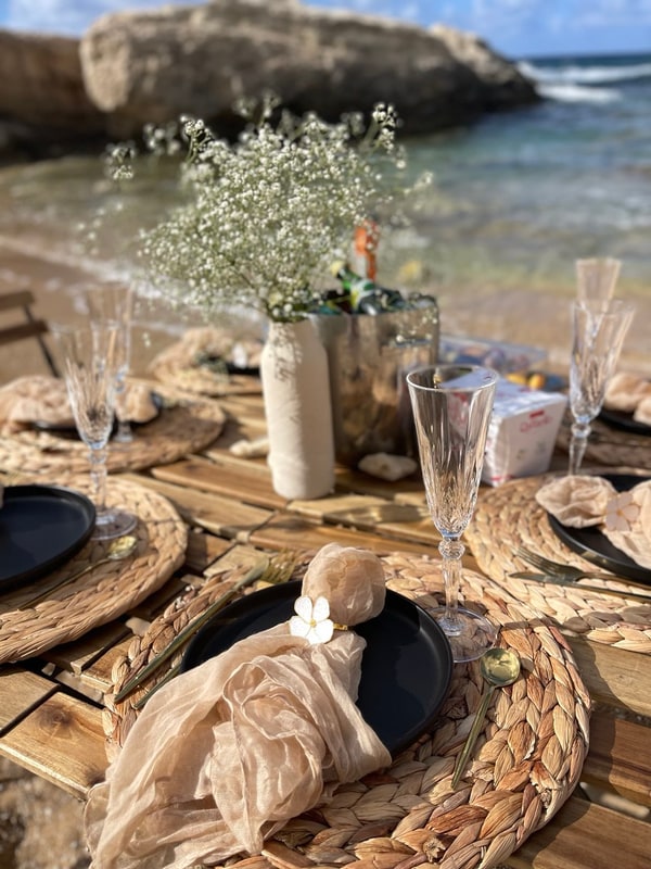 A picnic table with woven placemats and glassware beside the sea