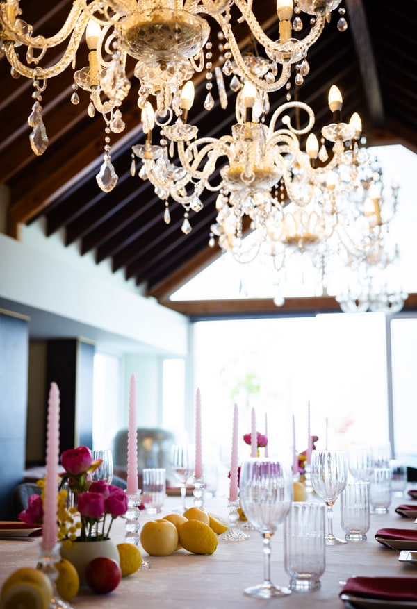 A long table with flowers, pink candles and lemons beneath chandeliers